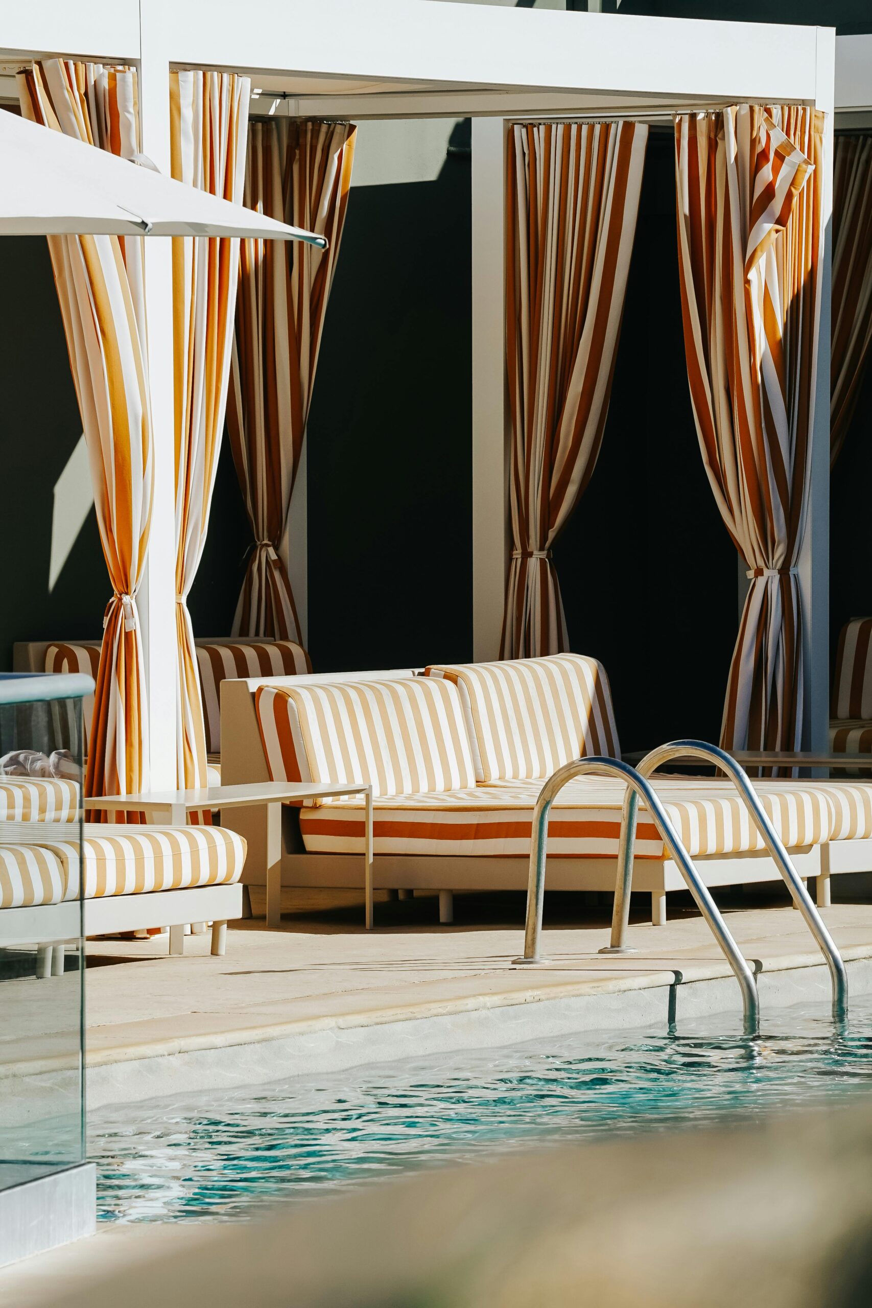 Sunny poolside cabana with striped curtains and loungers next to a swimming pool.